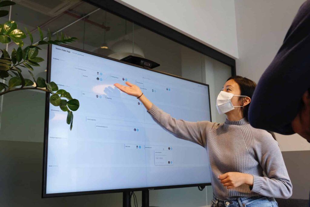 A woman wearing a face mask is pointing to a screen displaying web design.